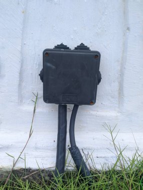 Beyaz bir duvara monte edilmiş siyah bir elektrik bağlantı kutusunun yakın çekimi. İki siyah kablo aşağıdan kutuya giriyor..