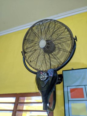 Parlak sarı bir duvara dayalı, metal kafesi ve kontrol düğmeleri olan antika bir siyah duvara monte edilmiş elektrikli vantilatör..