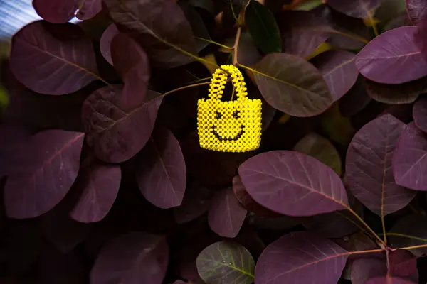 Canlı, sarı minyatür boncuklu bir çanta ve gülümseyen bir yüz mor yaprakların arka planında asılı duruyor. El sanatları, moda ve doğa ile ilgili konular için mükemmel. Yüksek kalite fotoğraf