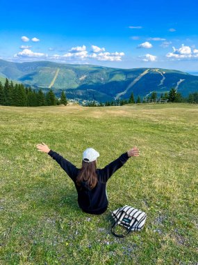 Siyah kapüşonlu ve şapkalı genç bir kadın yazın dağlarda oturur. Yakınlarda bir yerde bir yaz çantası yatıyor. Kadınlar geri döndü. Yüksek kalite fotoğraf