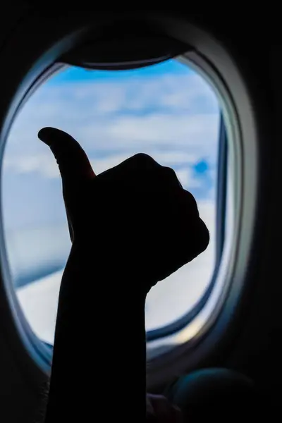 stock image A silhouette of a hand showing a thumbs-up gesture against the backdrop of a blue sky and clouds, seen through an airplane window. Low Key