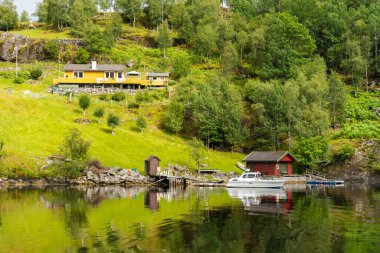 Bergen, Norveç - 11 Temmuz 2024: Bergen, Norveç yakınlarındaki fiyorttan bir gemi yolculuğunun görüntüsü, büyüleyici evler, yemyeşil alanlar ve sakin çevreyi yansıtan sakin sular..