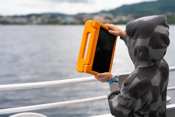 stock image Elementary boy tourist capturing scenic fjord views with an orange-cased tablet during a travel adventure. The child is wearing a hoodie and standing by the water, showcasing modern tourism in Norway