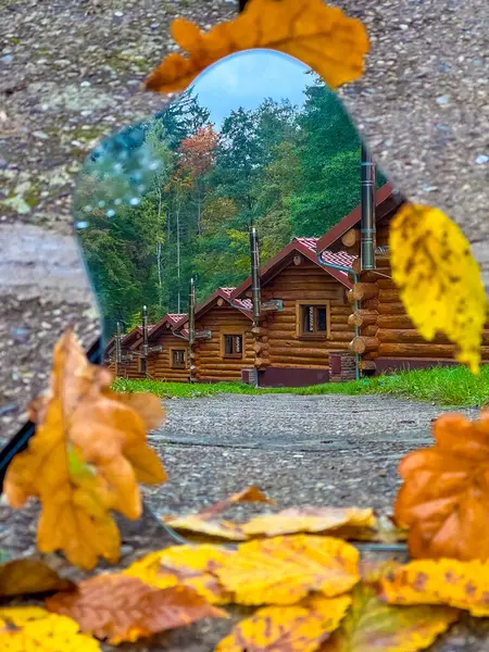 stock image A serene autumn scene in a Czech forest, capturing the reflection of rustic log cabins through a mirror framed by vibrant fallen leaves. The image evokes a peaceful, cottagecore atmosphere.