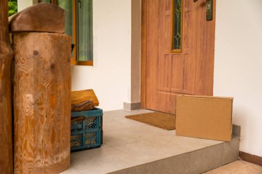 Bir yaz günü ahşap bir kır evinin girişine bırakılmış yeniden kullanılmış bir karton paket, beklenti ve paket almanın günlük rahatlığını simgeliyor. Çevrimiçi alışveriş kavramı.