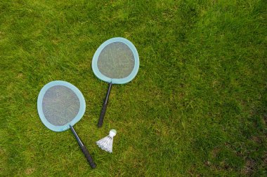 İki portakallı badminton raketi ve yeşil çimlerde dinlenen bir mekik horozu, yukarıdan çekildi. Açık havada spor yaparak geçirilen güneşli bir günün eğlencesini ve rahatlığını anımsatıyor. Boşluğu kopyala
