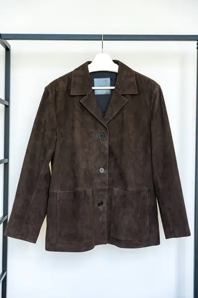 stock image A dark brown suede jacket with patch pockets hanging on a hanger against a white wall. This stylish outerwear piece represents a major fashion trend for autumn 2024 with its minimalistic design.