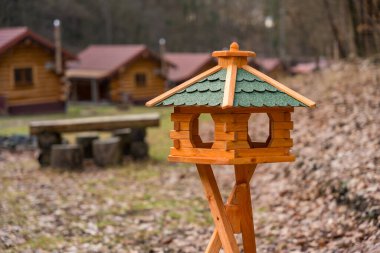 A close up wooden bird feeder stands in front of rustic log cabins in a forest setting in the Czech Republic. The earthy tones and natural surroundings evoke simplicity, and peacefulness in winter. clipart