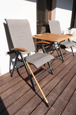 Yürüyen bir baston, ahşap bir masa ile güneşli ahşap bir güvertede, modern bir teras sandalyesine dayanır. Sahne, boş zamanların, ulaşılabilirliğin ve düşünceli tasarımın altını çiziyor. Sıcak tonlar ve insan yok..