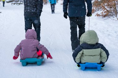Çek Cumhuriyeti, Spindleruv Mlyn 'de karlı bir yolda yetişkinler tarafından rehberlik edilirken iki çocuk kızağa biniyor. Kış manzarası, kar yağışı, aile etkileşimi. Göz hizasında çekilen dikiz aynası.