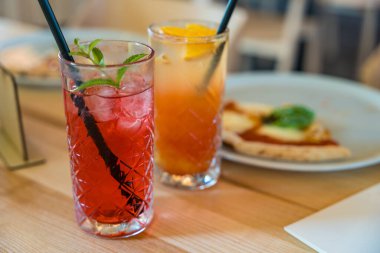 İki mocktail, nar şurubu ve kırmızı limonatalı Cinderella, yumuşak odaklı ahşap bir masada. Sıcak tonlar, yakın çekim görüntüsü ve sığ alan derinliği kopya alanı ile samimi bir ortam oluşturur.