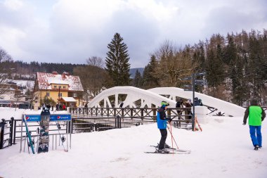 Spindleruv Mlyn, Çek Cumhuriyeti - 3 Ocak 2025: Karla kaplı bir yaya köprüsünün olduğu kış sahnesi. Kayakçılar ve turistler manzaralı dağlık kasabayı keşfediyorlar.