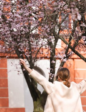 Bej ceketli beyaz bir kadın Prag 'daki turuncu bir binaya karşı çiçek açan kiraz çiçeklerinin altında kollarını kaldırıyor. Orta mesafeden, ılık tonlardan, Troja 'daki bahar atmosferinden yakalanmış.