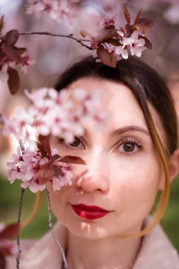 Prag, Troja Garden 'da kiraz çiçeklerinin çerçevelediği beyaz kadın. Yumuşak güneş ışığı yüzüne, kırmızı dudaklarına ve doğal güzelliğine vurgu yapıyor. Bulanık ön plan derinlik katıyor, rüya gibi, sanatsal bir hava yaratıyor..