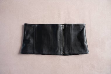 Ön fermuarlı siyah deri korse kemeri, yumuşak bej arka planda sergileniyor. Yapılandırılmış tasarım ve gösterişli doku onun şık ve keskin estetiğini vurguluyor..