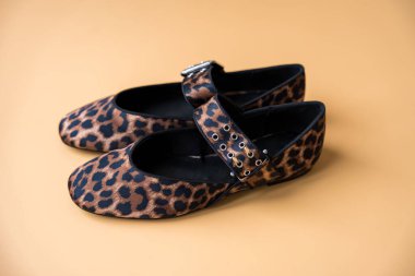 Bej arkaplana yerleştirilmiş şık tokalı bir çift leopar desenli tekstil bale ayakkabısı. Çekim yukarıdan aşağıya doğru çekiliyor, dokuyu vurguluyor.