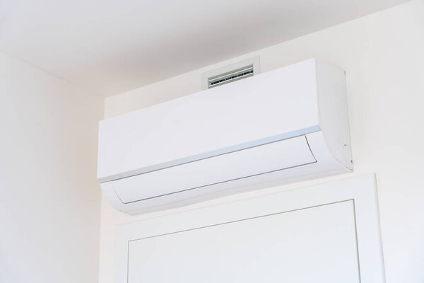 Close-up of a white air conditioner unit mounted on a white wall above a door in a modern home interior. Minimalist climate control technology for energy efficiency and comfort. No people, copy space.