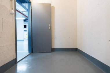 Modern bir yeraltı otoparkında, ağır metal kapısı olan güvenli bir yeraltı deposu. Sanayi bölgesinde beton zemin, beyaz duvarlar ve açıkta kalan borular yer alıyor..