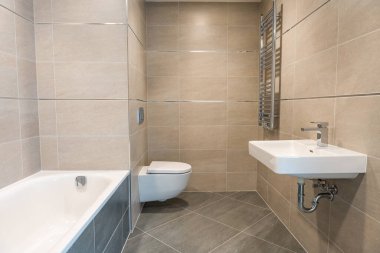 İçinde banyo küveti, duvara monte edilmiş lavabo ve bej seramik kiremitli tuvalet bulunan modern banyo tasarımı. Krom ısıtmalı havlu korkuluğu da dahil. Minimalist estetik, insan yok, kopyalama alanı.