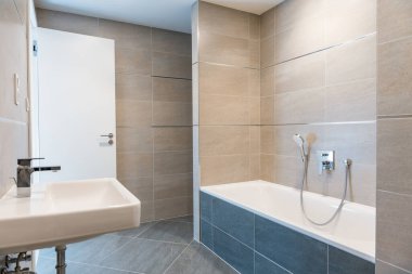 Minimalist banyo, beyaz küvet, duvara monte edilmiş lavabo ve krom musluk. Bej ve gri seramik fayanslar nötr ve zarif bir tasarım oluşturur. İnsan yok, kopyalama alanı.