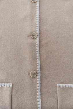 Bej renkli yün polyester bir ceketin yakın çekimi, dikey açı, yumuşak ışıklandırma, sığ derinlik, düğme sırasını gösteren ve beyaz işlemeli süsleme. Doku odaklı moda detayı, insan yok..