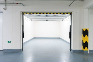 Çek konut binasındaki temiz, boş bir yeraltı garajının geniş açılı görüntüsü. Açık metal kapı, güvenlik işaretleri ve endüstriyel ışıklandırma. İnsan yok, boşluk kopyala, tarafsız ses tonları.