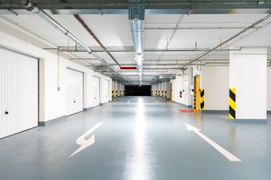 Temiz, modern bir yeraltı garajının geniş açılı görüntüsü. Kapalı depolama birimleri, yerde oklar ve parlak endüstriyel aydınlatma. Derin bir alan derinliği ile vurulmuş. İnsan yok, kopyalama alanı.