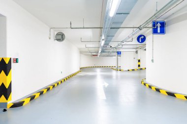 Kıvrımlı şeritli temiz yeraltı otoparkı, parlak ışıklandırma, sarı-siyah güvenlik işaretleri ve yön işaretleri. Geniş açı mercek, düşük perspektif, endüstriyel iç mekan. İnsan yok, kopyalama alanı.