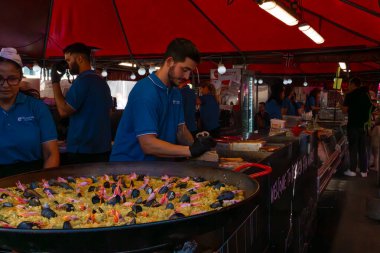Bergen, Norveç - 9 Temmuz 2024: Mavi üniformalı personel, Bergen ulusal balık pazarında kırmızı bir gölgelik altında büyük bir deniz ürünleri paella hazırlıyor