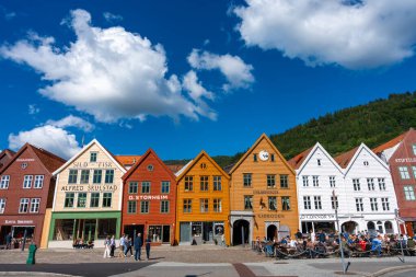 Bergen, Norveç - 9 Temmuz 2024: Açık mavi gökyüzü altında geleneksel Bryggen ahşap binalarının yakınındaki kalabalık. UNESCO miras bölgesinde tabelalar, kafeler ve sokak hayatı ile aydınlık cepheler.