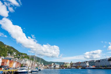 Bergen, Norveç - 9 Temmuz 2024: Mavi gökyüzü ve beyaz bulutlu güneşli bir günde, Bryggen limanındaki renkli rıhtım, yatlar ve tarihi binaların geniş açılı manzarası