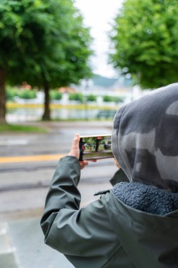 Norveç, Bergen 'de bir parkta kapüşonlu ceketli bir çocuk akıllı telefonuyla çekim yapıyor. Yaz yeşillikleri, günlük açık hava anı, içerik oluşturma, modern yaşam tarzı ve dijital alışkanlıklar.