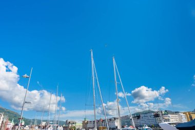 Bergen, Norveç - 9 Temmuz 2024: Uzun yelkenli direkleri, dağınık bulutlu parlak mavi gökyüzüne karşı limanın üzerinde yükselir. Şehir binaları, dağlar ve rıhtım manzarası marina ortamının altında görünüyor.