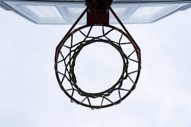 Bir basketbol potasının yakın plan görüntüsü. Altından dolanmış bir ağ, bulutlu gökyüzüne karşı yuvarlak çerçeveyi ve şeffaf seti gösteriyor. Spor ve minimalizmi vurguluyor.