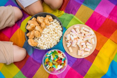 İki çocuk gökkuşağı battaniyesinde patlamış mısır, kruvasan, şekerleme ve şekerleme kaselerinde oturarak yaz pikniğinin tadını çıkarıyor.