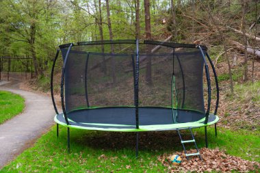 Orman parkında yeşil çimlerin üzerinde duran güvenlik duvarı ve merdiveni olan büyük bir trambolin. Top yakında. Baharın başında. Eğlence için mükemmel, açık hava eğlencesi, sağlıklı yaşam tarzı, aktif çocukluk..