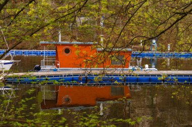 Sakin bir marinaya kenetlenmiş penceresi olan modern turuncu yüzen ev suya yansıyor. Ağaç dalları tarafından çerçevelenmiş bu sakin nehir kenarı manzarası eko-yaşam ve bahar sükunetini yakalıyor..