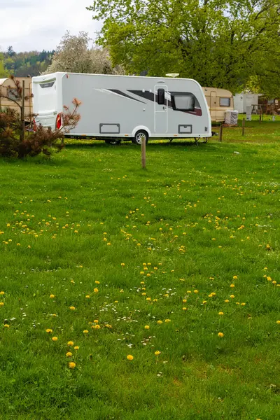 Çek Cumhuriyeti 'nde sakin bir bahar kampında beyaz, modern bir karavan. Yeşiller yemyeşil çimenler, çiçek açan karahindibalar ve ağaçlar dışarıda huzurlu bir yolculuk ortamı yaratır. Görünürde kimse yok, boşluğu kopyala