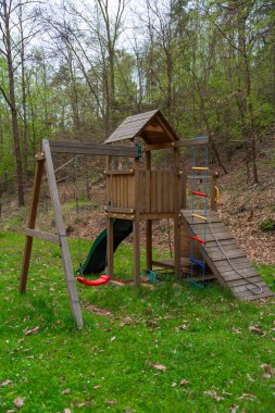 Açık hava ahşap oyun yapısı, kırmızı salıncak, yeşil kaydırak ve tırmanma ipleri orman ağaçlarının yanındaki çimlerin üzerinde duruyor. Kırsal alan, erken bahar yaprakları. Doğa oyunları, aile, çocukluk temaları için ideal.