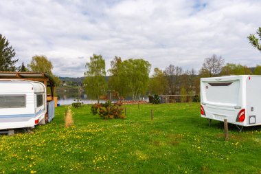 Vintage camper trailers parked by a quiet lakeside camping area in spring. Green grass, wildflowers, wooden shelter and peaceful outdoor travel lifestyle. No people, copy space