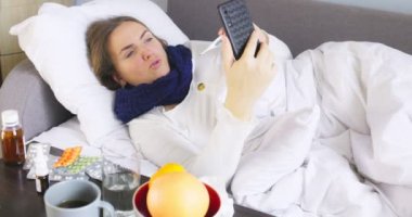 Sick young woman with bad cold, flu or coronavirus fever holding thermometer while consulting a doctor by video link via a smartphone. Sick woman having online medical consultation video call