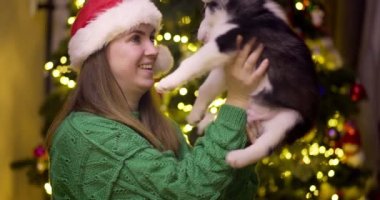 Güzel bir kadın Noel ağacının yanında otururken köpek yavrusuyla oynayıp eğleniyor. Genç bayan kırmızı Noel Baba şapkasıyla köpeğine sarılıyor.