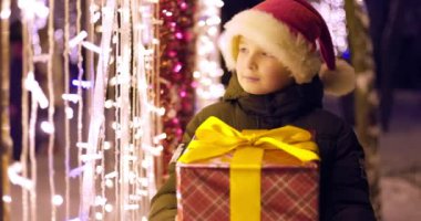 Noel Baba şapkalı çocuk Noel ışıklarının yanında hediye kutusuyla. Noel hediyeleri, tatil atmosferi. Noel ve Yeni Yıl konsepti. Yüksek kalite 4k görüntü