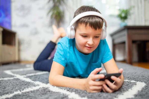 A child boy with phone and headphones is lying on the floor watching cartoons, listening to music, playing games, talking on the phone