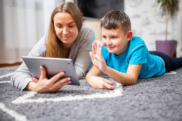 Happy mother and kid son greeting online looking at web camera using tablet pc for video call lying on carpet. Smiling mom and child having fun talking in video chat
