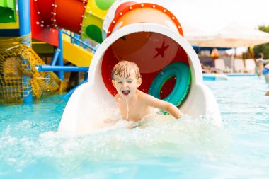 Happy laughing little boy playing on water slide in outdoor swimming pool on a hot summer day. Child sliding on aqua playground in tropical resort clipart