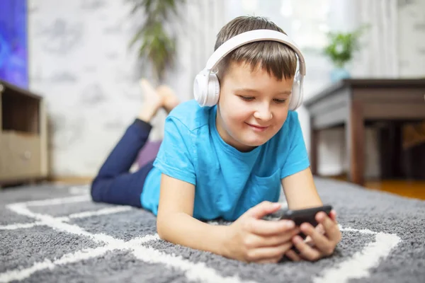 A child boy with phone and headphones is lying on the floor watching cartoons, listening to music, playing games, talking on the phone
