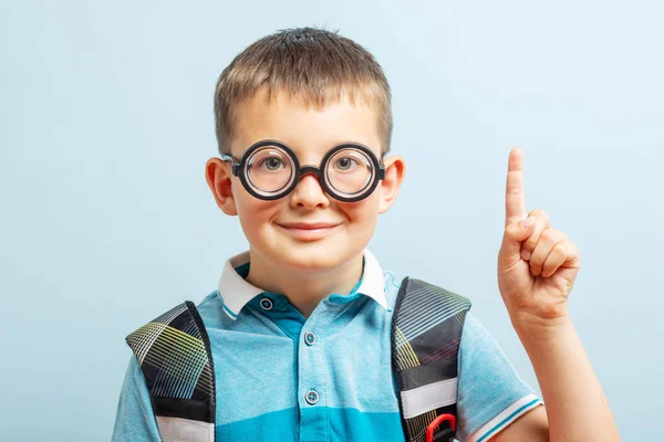 A cute school boy in glasses with raised index finger on blue background. The child points the finger up. Back to school