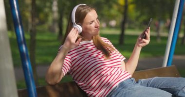 Kablosuz kulaklıklı genç bir kadın salıncakta sallanıyor, şehir parkında müzik dinliyor. Yüksek kalite 4k görüntü