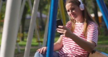 Kablosuz kulaklıklı genç bir kadın salıncakta sallanıyor, şehir parkında müzik dinliyor. Yüksek kalite 4k görüntü